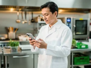 Voedselveiligheidsinspecteur in witte jas controleert digitale checklist op smartphone in professionele commerciële keuken.