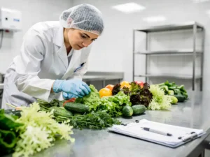 Voedselveiligheidsinspecteur in witte jas onderzoekt verse groenten op roestvrijstalen keukentafel met klembord nearby.