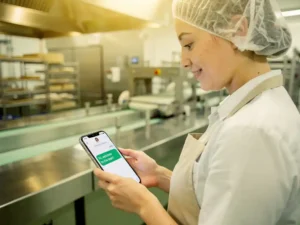Nieuwe medewerker in haarnetten schort leest WhatsApp-bericht naast roestvrijstalen voedsellijn tijdens onboarding.